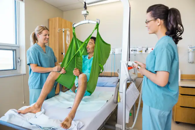 Female nurses looking at mature male patient sitting on cloth of hydraulic lift over bed at hospital