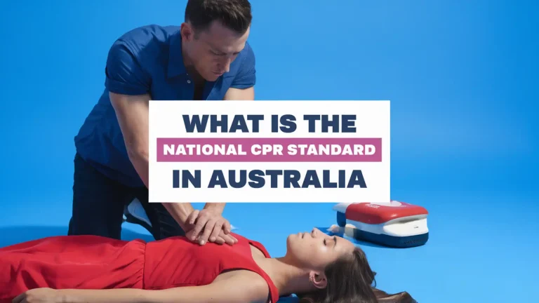 The image shows a man performing CPR on a person lying on their back, set against a bright blue background. The person receiving CPR is wearing a red dress, and there is a defibrillator placed next to them. Overlaying the image is a bold text that reads: "What is the National CPR Standard in Australia", with "National CPR Standard" highlighted in a pink band.